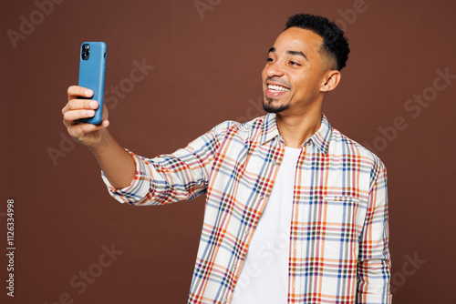 Young man of African American ethnicity wear blue shirt casual clothes doing selfie shot on mobile cell phone post photo on social network isolated on plain brown background studio. Lifestyle concept.