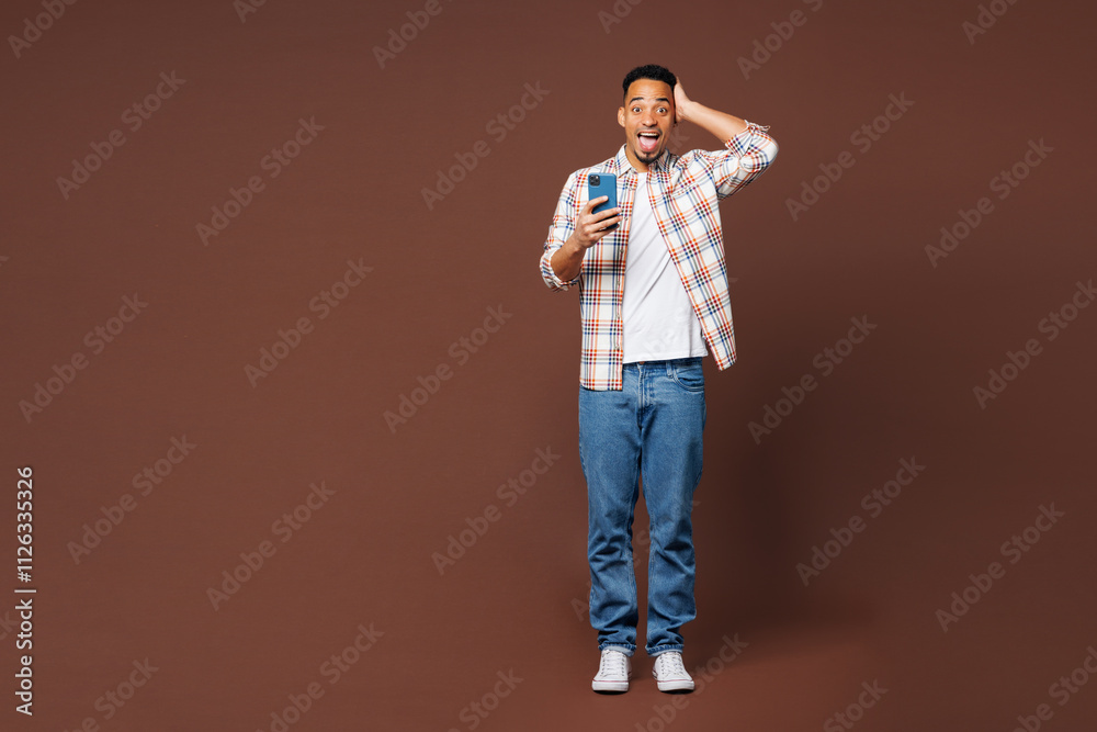 Full body shocked surprised young man of African American ethnicity he wear blue shirt casual clothes hold in hand use mobile cell phone isolated on plain brown background studio. Lifestyle concept.