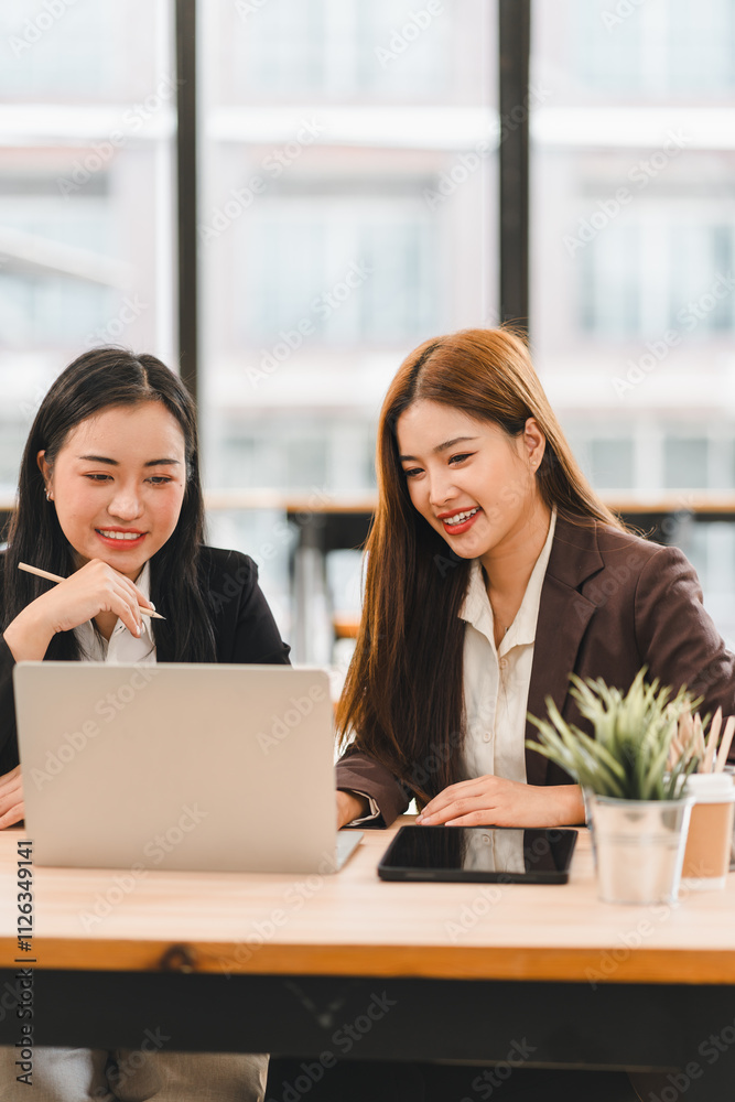 Two businesswomen collaborating on project while working on laptop in modern office. They are smiling and engaged, with tablet and potted plant on desk Two businesswomen collaborating on project while working on laptop in modern office. They are smiling and engaged, with tablet and potted plant on desk