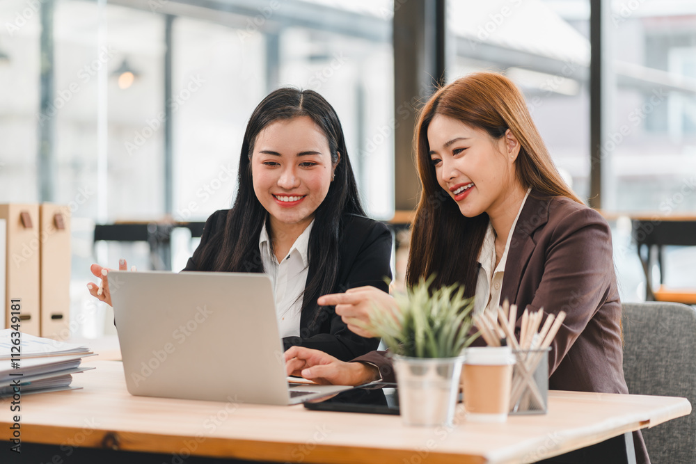 Two businesswomen in formal attire smiling and collaborating on laptop in modern office, with small plant and coffee cup on desk, showcasing teamwork and productivity Two businesswomen in formal attire smiling and collaborating on laptop in modern office, with small plant and coffee cup on desk, showcasing teamwork and productivity