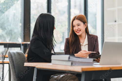 Two businesswomen discussing work at modern office desk with laptop, documents, and books, sharing ideas and smiling in collaborative and professional