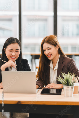 Two businesswomen collaborating on project while working on laptop in modern office. They are smiling and engaged, with tablet and potted plant on desk