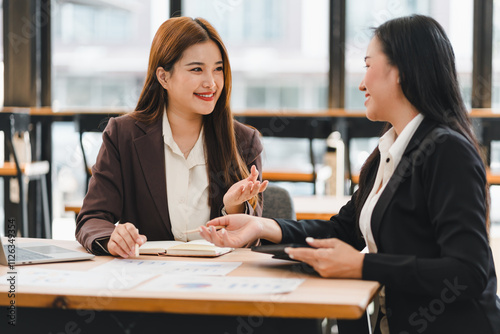 Two businesswomen in formal attire discussing work at desk with documents and laptop, smiling and engaging in professional conversation in modern office