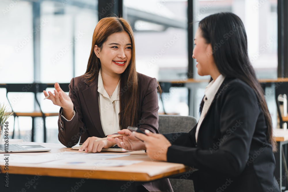 Two businesswomen in formal attire having cheerful discussion at modern office table, with documents and laptop in front of them, showcasing teamwork and collaboration Two businesswomen in formal attire having cheerful discussion at modern office table, with documents and laptop in front of them, showcasing teamwork and collaboration