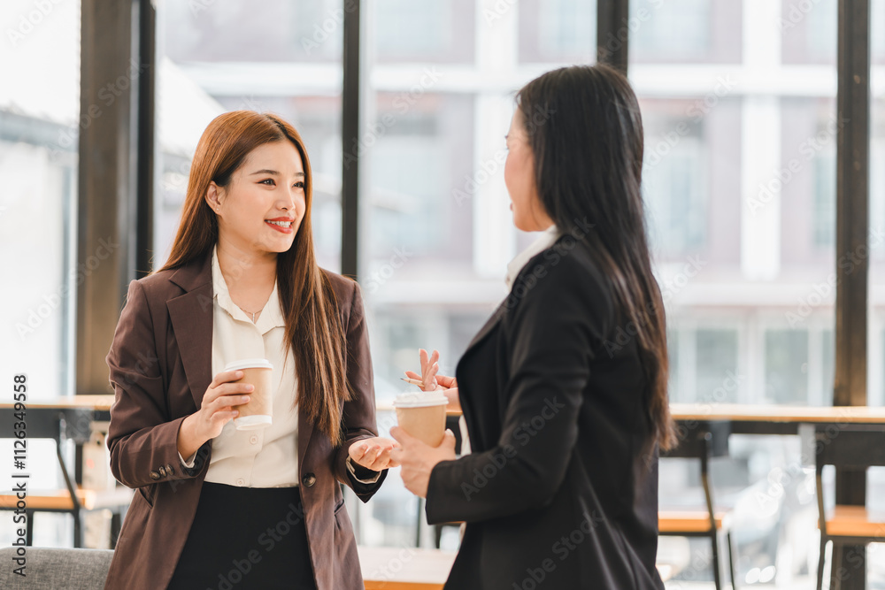 Two professional women in business attire having friendly conversation while holding coffee cups in modern office with large windows Two professional women in business attire having friendly conversation while holding coffee cups in modern office with large windows