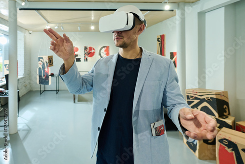 Young man in virtual reality helmet scrolling through extended artworks while standing in gallery of modern arts photo