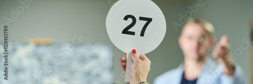 Banner of hand of young female participant of auction raising paddle with number twenty seven in front of camera photo