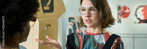 Banner of young brunette woman in eyeglasses explaining her opinion about represented artworks to African American visitor