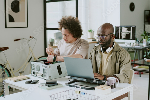 African American man using laptop on cooling pad to code while his biracial colleague repairing computer near him