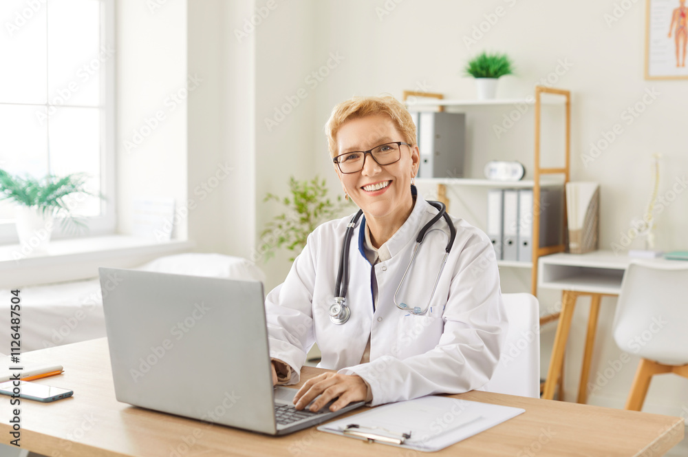 Portrait of a mature senior happy smiling female doctor sitting at the desk working in medical office and looking cheerful at camera. Woman physician wearing stethoscope in clinic using laptop Portrait of a mature senior happy smiling female doctor sitting at the desk working in medical office and looking cheerful at camera. Woman physician wearing stethoscope in clinic using laptop