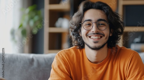 Lifestyle portrait of happy gamer, ethnic features, trendy eyewear, dark flowing hair, vibrant orange shirt, comfortable home setting, grey couch detail, natural window light, minimalist room design,