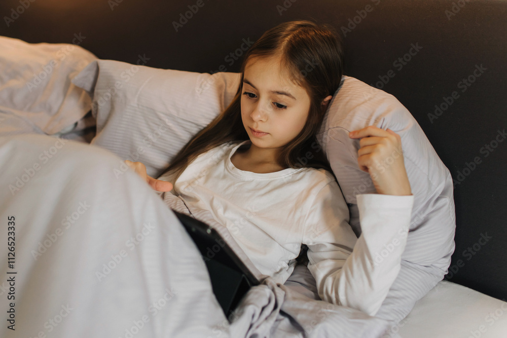 Pretty teenage girl using app on tablet, talking on online video call, laughing, secretly watching movie, playing online games, lying under covers on bed. Generation alpha concept Pretty teenage girl using app on tablet, talking on online video call, laughing, secretly watching movie, playing online games, lying under covers on bed. Generation alpha concept