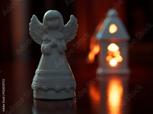 
The photo shows a white ceramic angel figurine in the foreground, with warm, soft lighting. In the blurred background, a glowing lantern creates a cozy, festive atmosphere. The focus emphasizes the 
