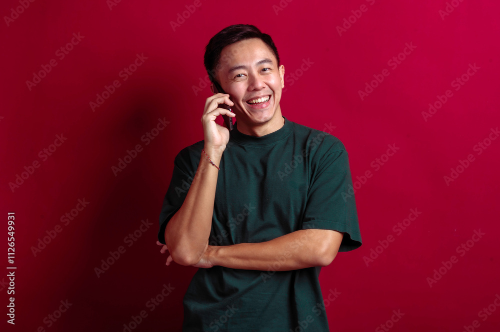 Asian man in a green T-shirt smiling warmly while talking on a smartphone, arms crossed casually, standing against a vibrant red background, conveying a sense of joy and connection