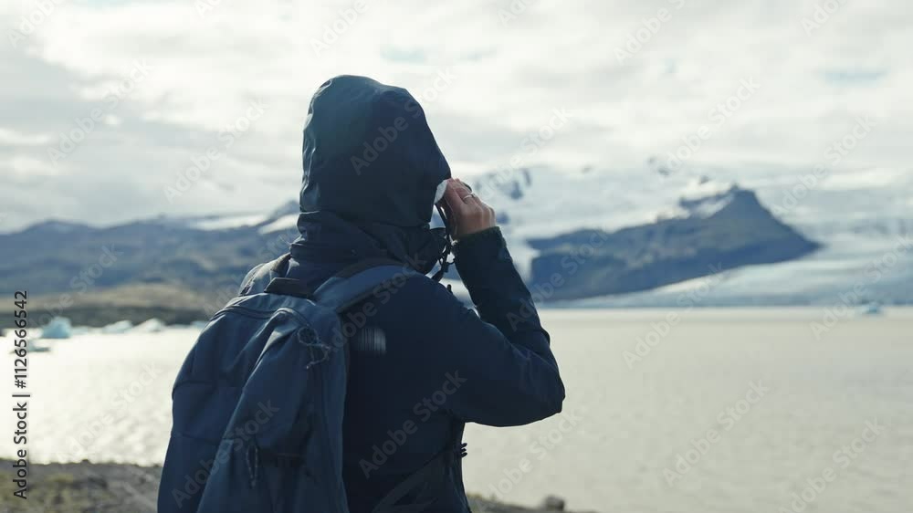 Middle eastern person in a blue hoody and backpack looking in binoculars at Jokulsarlon lake