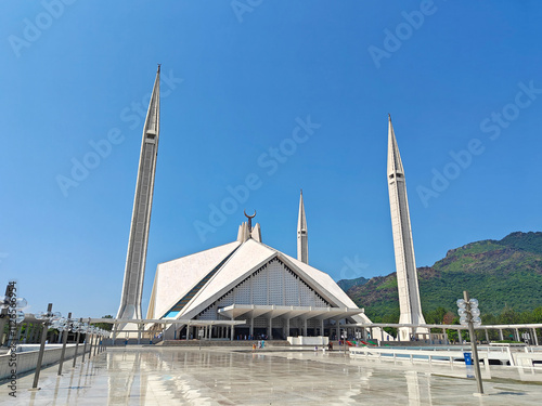 Shah Faisal Mosque in Islamabad with its iconic geometric design, tall minarets, and a vast courtyard under a clear blue sky, set against the backdrop of green Margalla Hills on a sunny day