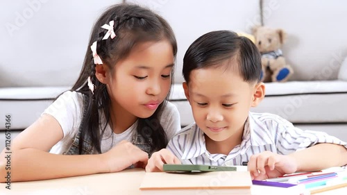 Asian elder sister and younger brother using mobile phones to watch movies or cartoons in living room. They enjoying and having fun with entertainment on phones, using home internet