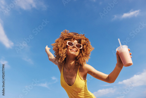 Joyful woman with curly hair enjoying a drink against a clear blue sky