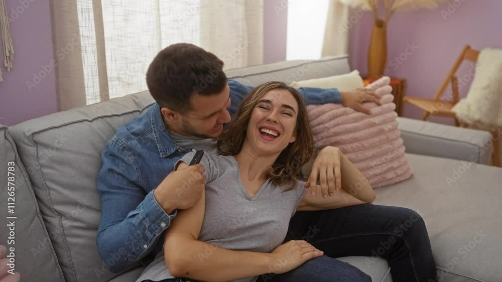 Couple watching television in cozy living room smiling happily on comfortable couch inside their home apartment Couple watching television in cozy living room smiling happily on comfortable couch inside their home apartment