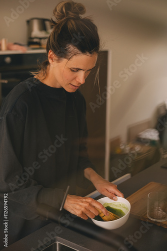 Woman cook matcha tea latte at home with bowl and bamboo whist. Happy girl with high bun mix matcha powder in bowl in the kitchen. photo