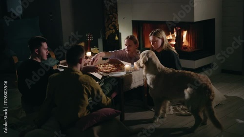 A group of young people enjoying pizza at a table near a glowing fireplace with a playful Golden Retriever beside them, sharing warm moments before Christmas Eve.