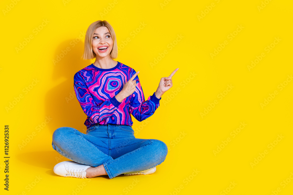 Full size photo of pretty young girl look point empty space sit floor wear trendy colorful blue outfit isolated on yellow color background Full size photo of pretty young girl look point empty space sit floor wear trendy colorful blue outfit isolated on yellow color background