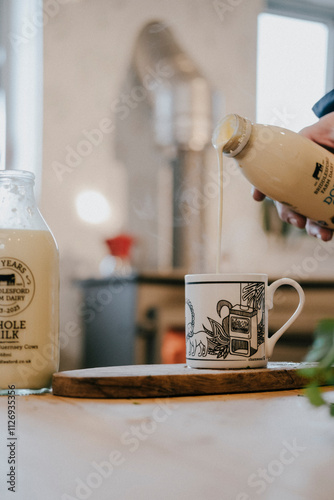 A person is pouring milk into a cup. The cup is white and has a picture of a house on it. The milk is coming from a bottle that says 