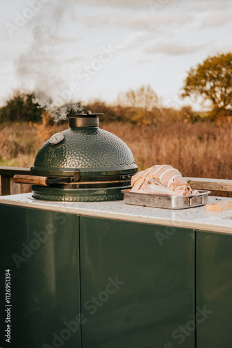 A green ceramic grill sits on a counter next to a tray of meat. The grill is emitting smoke, indicating that it is in use. The scene has a rustic, outdoor feel, with the grill photo