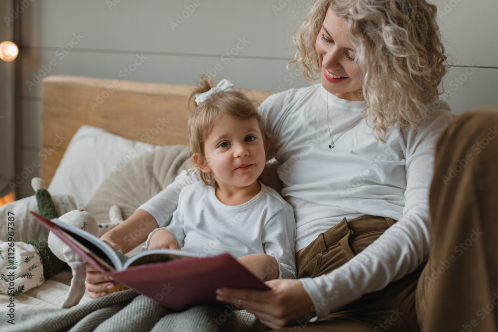 Close-up of a smiling woman sitting on a bed reading a book with her young daughter