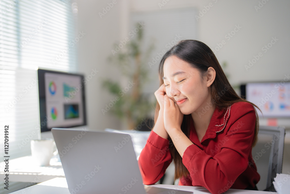 Young office worker taking a break from work, holding hands near her face and smiling, enjoying her achievements, feeling satisfied with work done, sitting at desk with laptop and desktop computer Young office worker taking a break from work, holding hands near her face and smiling, enjoying her achievements, feeling satisfied with work done, sitting at desk with laptop and desktop computer