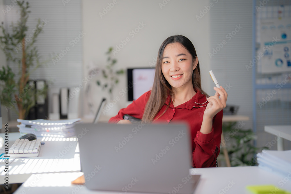 Young asian businesswoman holding a pen and smiling while working with a laptop in a modern office, showcasing confidence and professionalism in the workplace Young asian businesswoman holding a pen and smiling while working with a laptop in a modern office, showcasing confidence and professionalism in the workplace