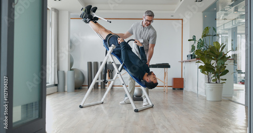 People, inversion table and upside down for physical therapy with support, balance and recovery at clinic. Physiotherapist, men and patient with fitness bench, biokinetics and rehabilitation process photo