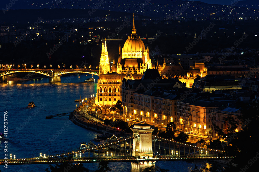 Fototapeta premium Stunning night view of Budapest featuring the illuminated Parliament building, Chain Bridge, and Danube River with reflections and boats. A vibrant cityscape highlighting Hungarian landmarks and archi