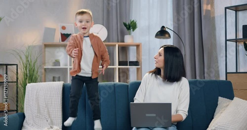 Woman admiring of her cute 2-aged son which jumping near her on couch and then hugging him photo