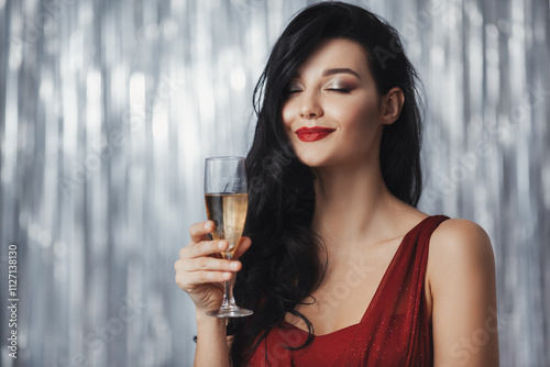 Принти на полотні Beautiful brunette woman in a red dress holding a champagne glass with a serene smile, set against a shimmering silver background