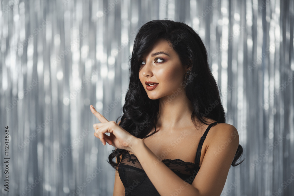 Obraz premium Beautiful brunette woman wearing black lingerie and pointing to the side with a sultry expression, set against a glittering silver backdrop. Perfect for luxury, fashion, and promo themes. 