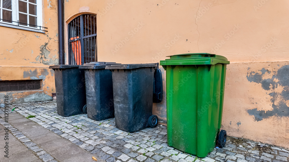 Four trash bins, including a green recycling bin, lined up against a weathered wall, highlighting urban waste management challenges Four trash bins, including a green recycling bin, lined up against a weathered wall, highlighting urban waste management challenges