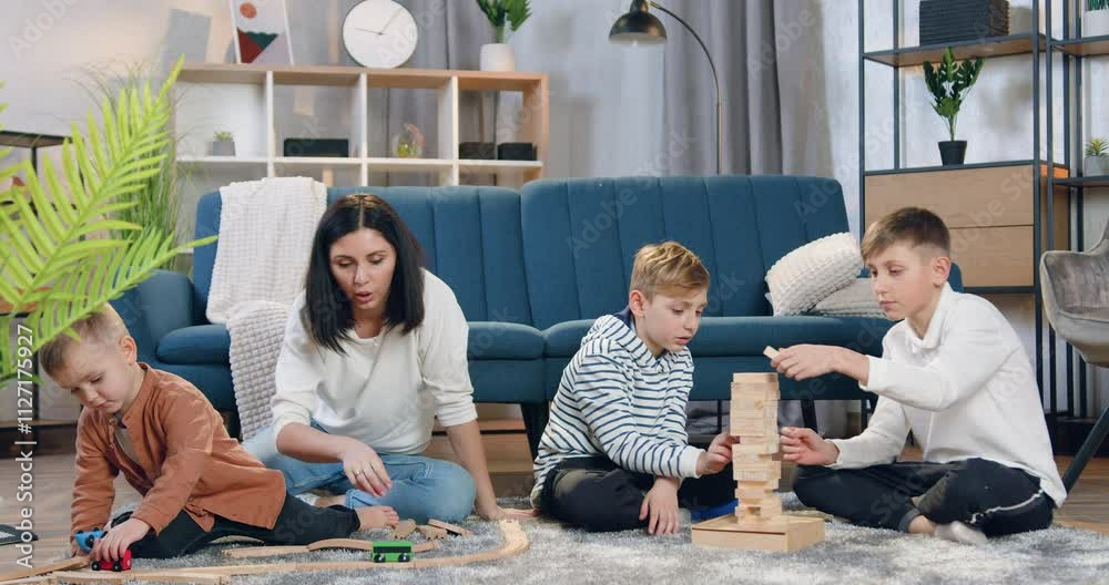 Boys playing with wooden blocks and demolish the built tower while their loving pretty mother playing with their smallest brother with car on car track