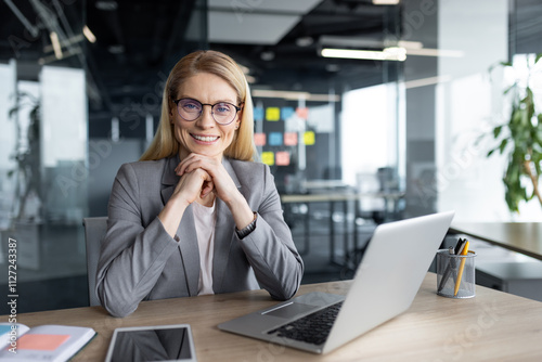 Wallpaper Mural A confident businesswoman in a professional setting, sitting at her desk with a laptop and smartphone, smiling at the camera, symbolizing success and professionalism in the workplace. Torontodigital.ca