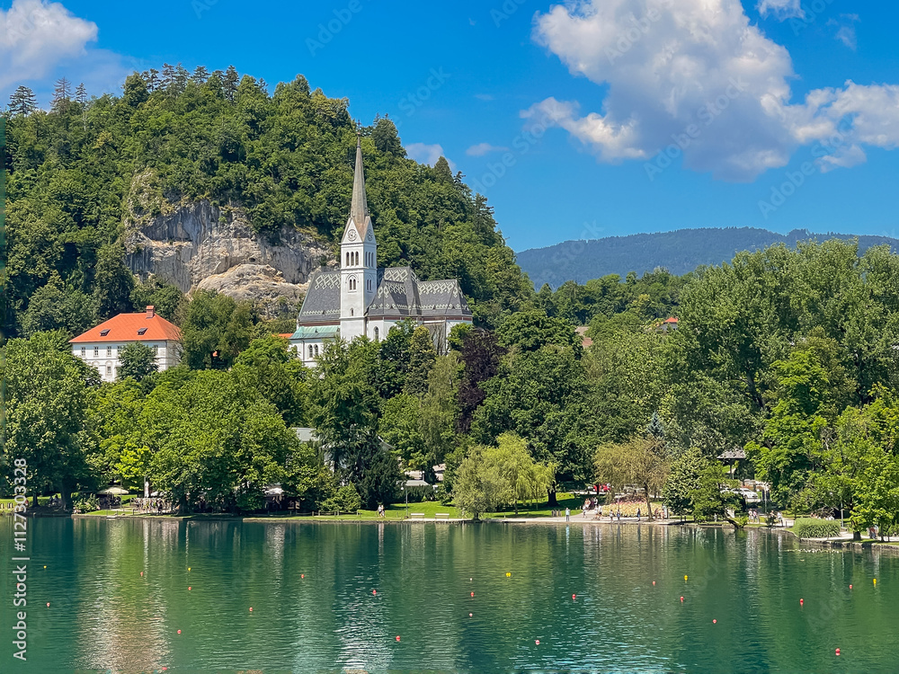 Fototapeta premium Lake Bled, Slovenia - June 28, 2024: St. Martin Church with gray-white mosaic roof surrounded by green foliage under blue cloudscape. White cliffs in back. Large white parish residential and office ho