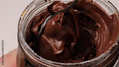 spoon scooping nut paste from glass jar. Melted chocolate nut nougat drips from above, creating. Creamy texture and glossy finish of the chocolate dessert.  