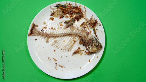 Fish Bones on The White Plate and Green Background. The Food Waste. Remains of Fried Fish. Fishbone and Leftover Meat of Crucian Carp After Eat