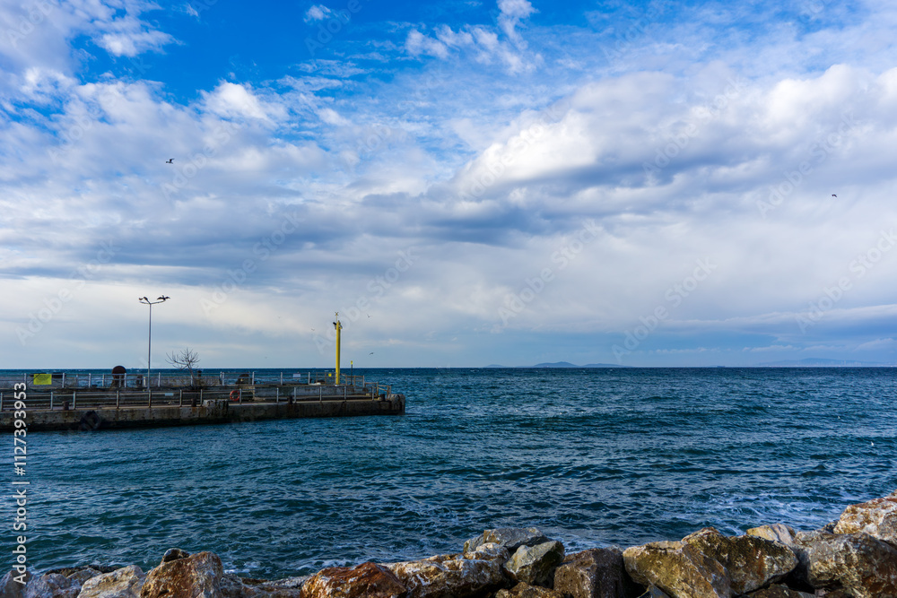 Rocky Coastline with Jetty and Cloudy Sky. Yellow navigation light post or Lighthouse on the concrete pier. Serene Waterfront View with a Rocky Foreground Rocky Coastline with Jetty and Cloudy Sky. Yellow navigation light post or Lighthouse on the concrete pier. Serene Waterfront View with a Rocky Foreground
