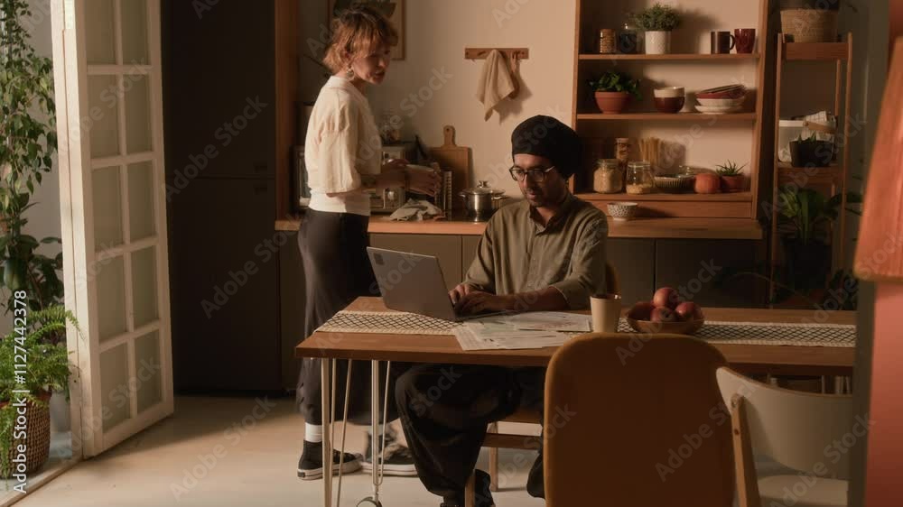 Full shot of young adult Caucasian wife with mug coming up to Indian husband in turban who sitting at dining table with laptop and paying utility bills in domestic kitchen Full shot of young adult Caucasian wife with mug coming up to Indian husband in turban who sitting at dining table with laptop and paying utility bills in domestic kitchen