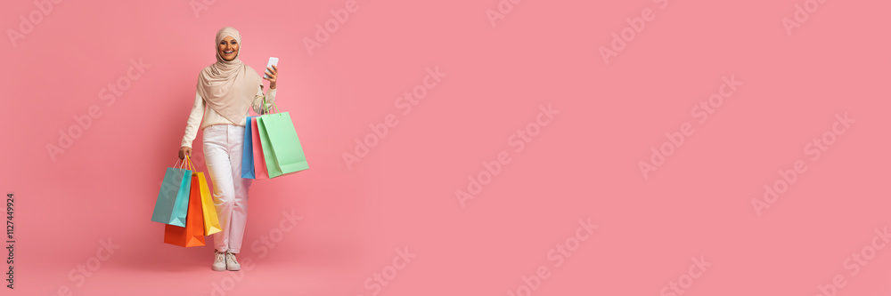 Muslim woman dressed in casual attire holds multiple shopping bags while smiling happily. She enjoys her shopping experience during a sale, with a bright pink background enhancing the festive mood.