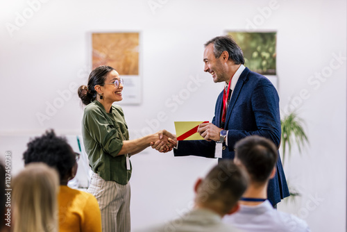 Front view of businesswoman receiving award from businessman in front of business professionals applauding at business seminar in office building