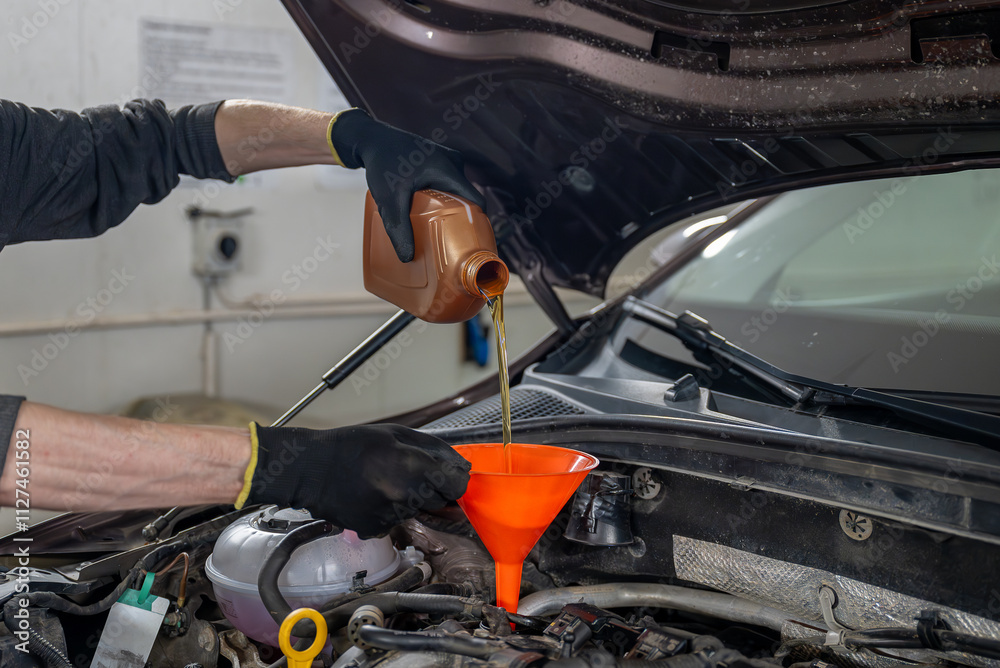 Fototapeta premium Mechanic in gloves pouring motor oil into a car engine using an orange funnel, in a workshop. Concept of vehicle maintenance 