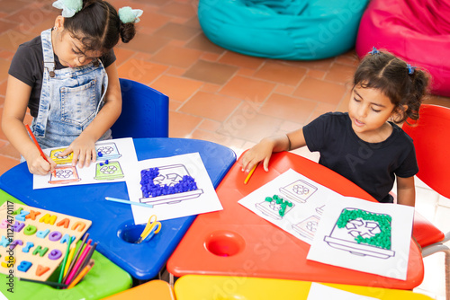 Dos niñas latinas de primaria realizando trabajos sobre el reciclaje en el aula de clases, con mesas coloridas