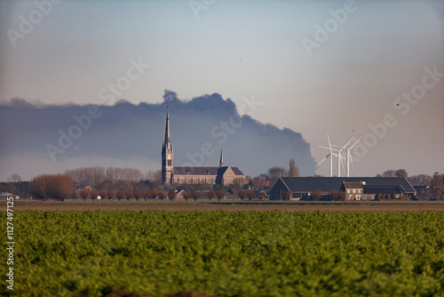 Large plume of smoke hangs over rural Dutch landscape, obscuring church and farm buildings. photo