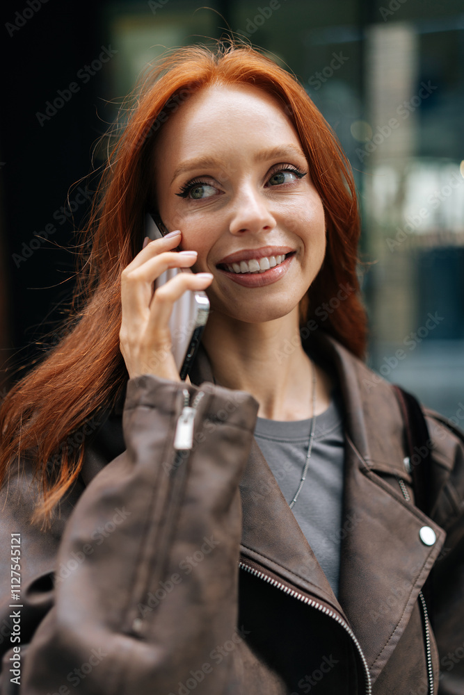 Closeup vertical portrait of redhead woman talking on phone while walking in city on cloudy day. Happy ginger female enjoying cheerful phone conversation. Cute joyful lady enjoying casual day out. Closeup vertical portrait of redhead woman talking on phone while walking in city on cloudy day. Happy ginger female enjoying cheerful phone conversation. Cute joyful lady enjoying casual day out.